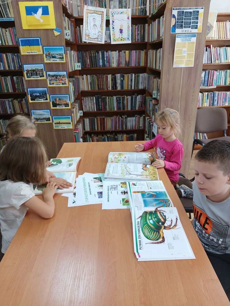 Czworo dzieci siedzi przy stole i ogląda książki o owadach. W tle regały z książkami.