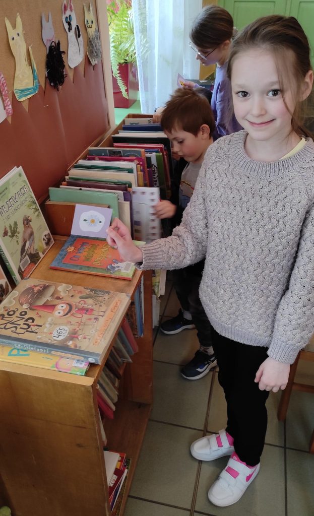 Uśmiechnięta dziewczynka w jasnym sweterku trzyma w ręce obrazek z białym kwiatkiem. W tle stoi dziewczynka i chłopiec oglądający książki stojące na regale.