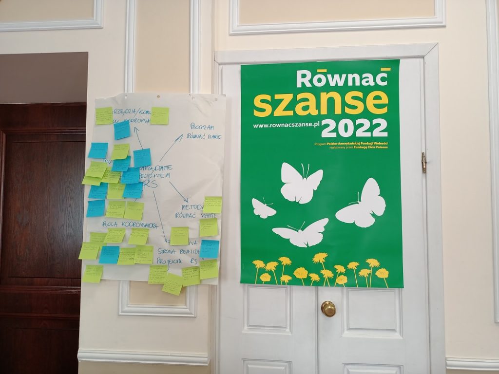 Dwa plakaty informacyjne zawieszone na ścianie i drzwiach. Z lewej strony plakat dotyczący zarządzania projektem RS. Z prawej plakat z napisem Równać Szanse 2022.