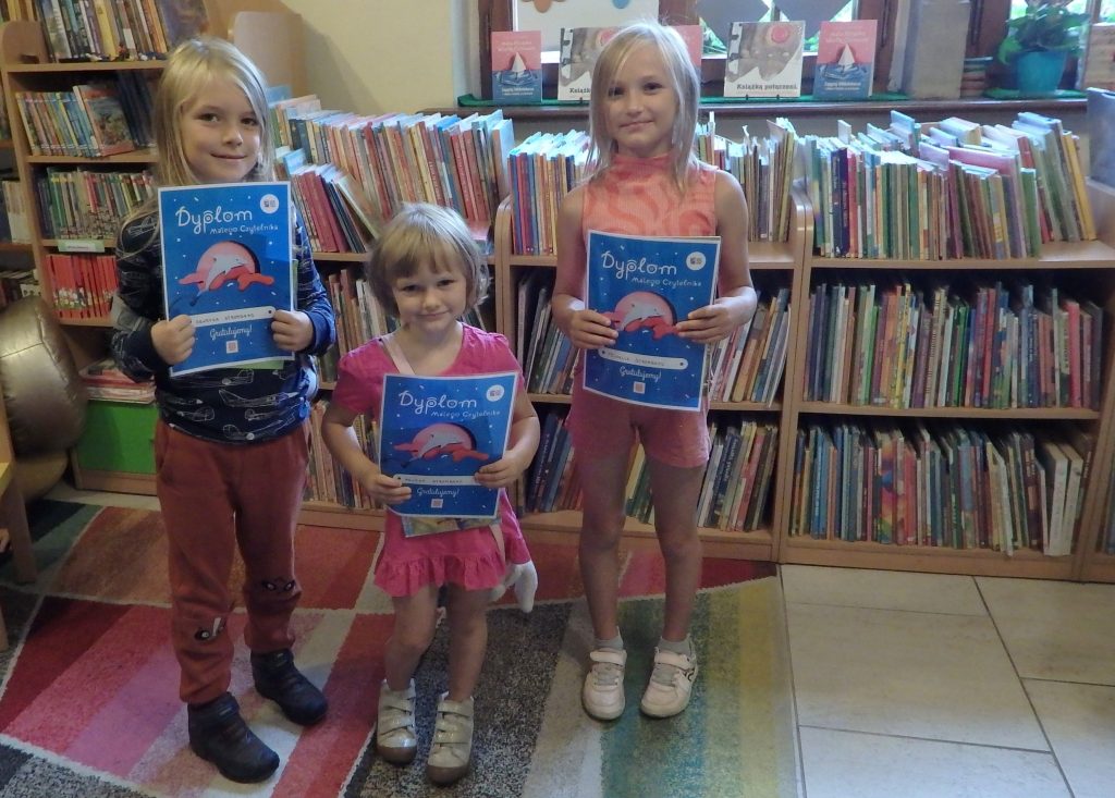 Na zdjęciu stoi troje dzieci, w rękach trzymają kolorowe dyplomy. W tle regały z książkami.