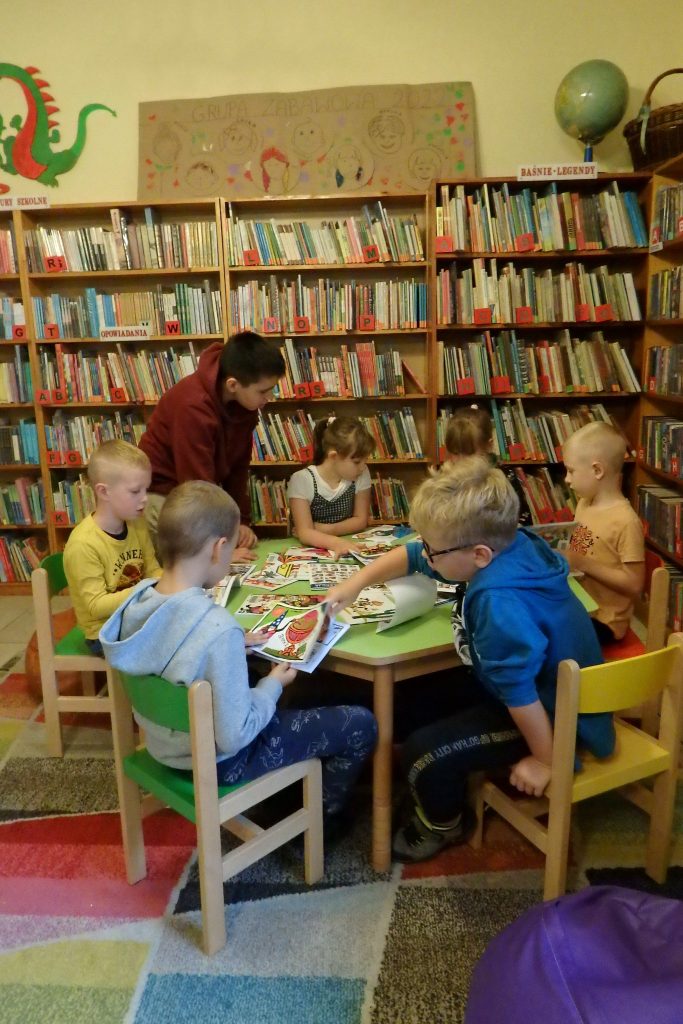 Na zielonym stoliku leżą komiksy „Tytus, Romek i A’Tomek”. Wokół stolika siedzą dzieci na kolorowych krzesełkach i przeglądają komiksy. W tle stoją regały z książkami.