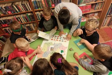 Przy stoliku siedzi grupka dzieci. Na środku stołu leży mapa Polski .Dzieci przeglądają książki. W tle regały z książkami.