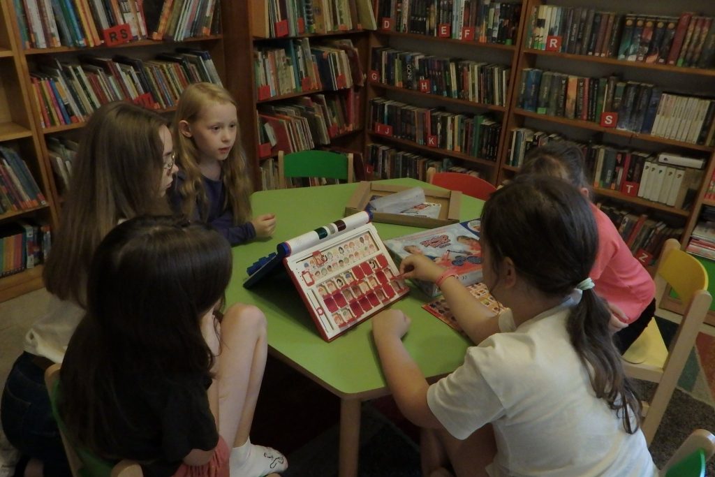 Przy stoliku siedzą dziewczynki, mają przed sobą grę ‘’Zgadnij kto to?’’. W tle regały z książkami.