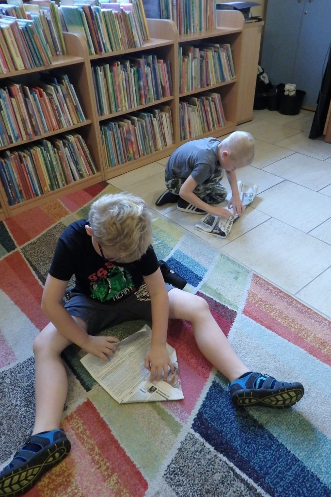Na kolorowym dywanie siedzi dwóch chłopców. Każdy z nich ma kartkę szarej gazety i składa ją tak by wyszedł samolot. W tle regały z książkami.