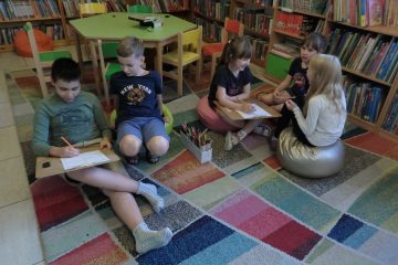 Na kolorowym dywanie po prawej stronie siedzą dziewczynki, a po lewej siedzą chłopcy. Każda z grup ma podkładkę z kartką na której malują. Na dywanie jest pudełko z kolorowymi kredkami. W tle zielony stolik, kolorowe krzesełka i regały z książkami.