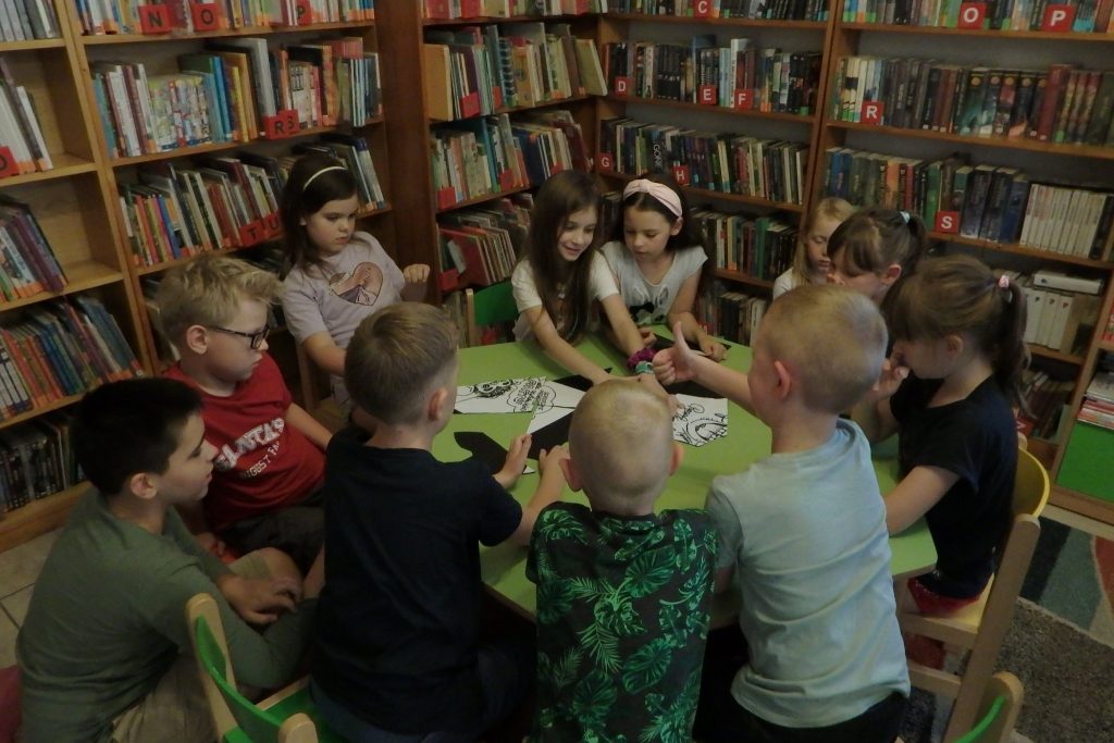 Na zdjęciu przy stoliku siedzi grupka dzieci. Mają elementy obrazka i próbują go ułożyć. W tle regały z książkami.