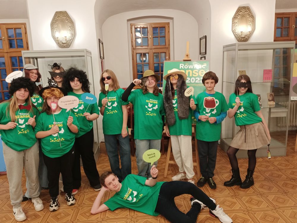 Grupa młodzieży stojąca w rzędzie, ubrana w zielone koszulki. Przed nimi leżący chłopiec.