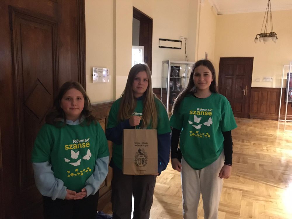 Trzy stojące dziewczynki ubrane w zielone koszulki. Środkowa dziewczynka trzyma w dłoniach papierową torebkę z napisem „Gmina i miasto Lwówek Śląski”.