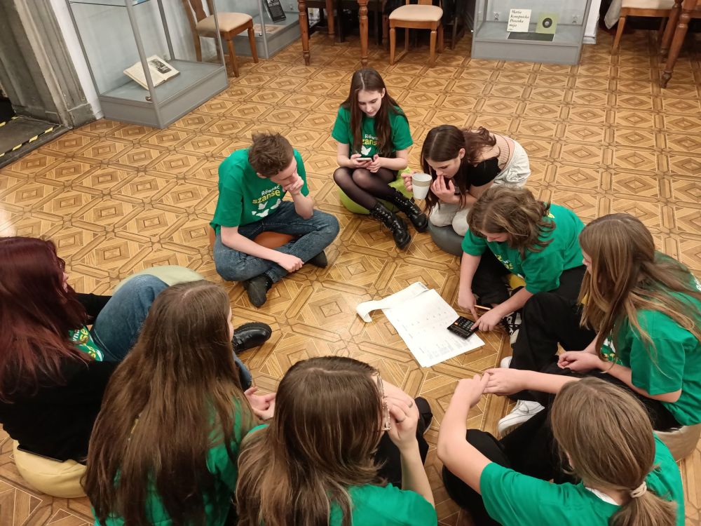 Grupa młodzieży siedząca w kole na podłodze. Przed nimi kartki i kalkulator.