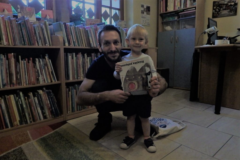Na zdjęciu stoi chłopczyk, w rękach trzyma książkę. Obok niego kuca jego tata. W tle regały z książkami, szafa oraz biurko pani bibliotekarki.