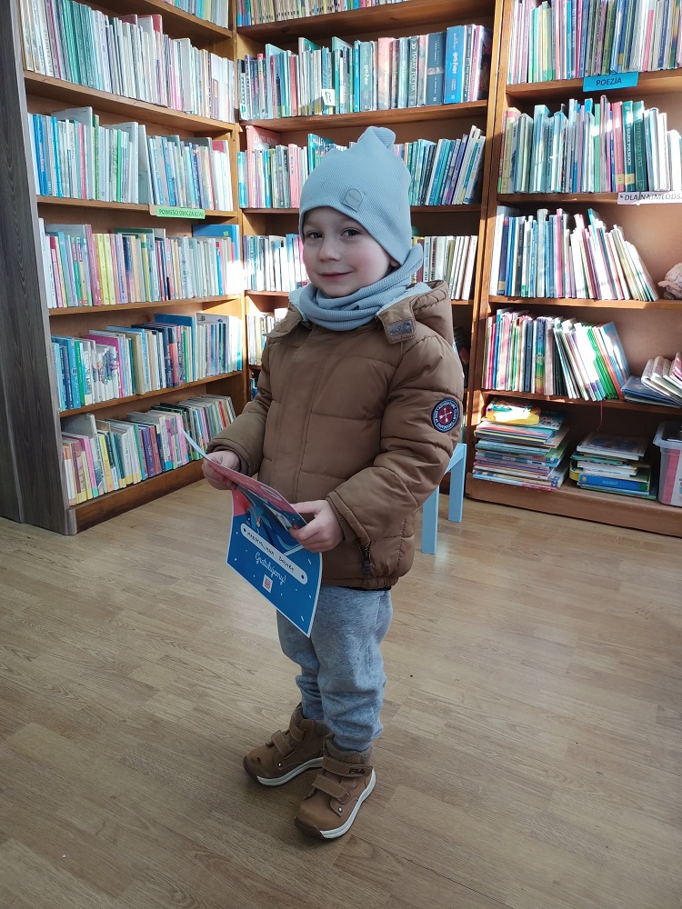 Stojący mały chłopiec, trzymający dyplom. W tle regały z książkami.