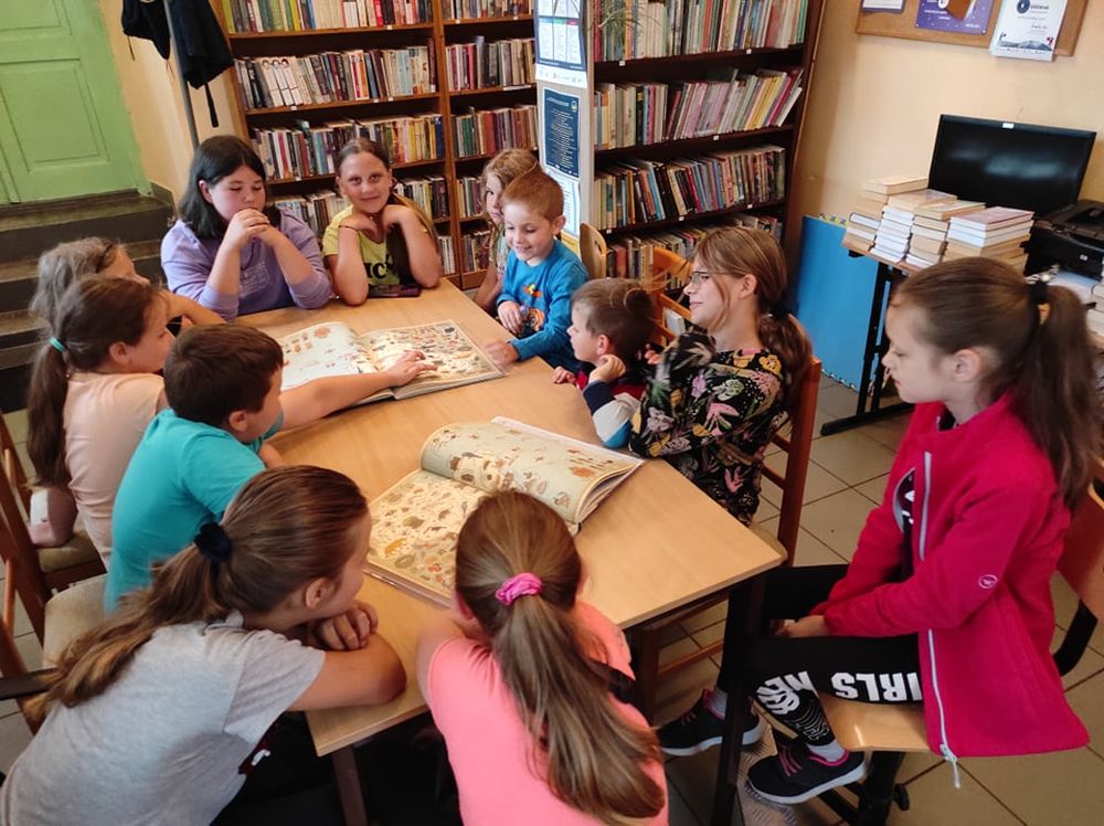 Dwanaścioro dzieci siedzi na krzesłach dookoła stołu. Na stole leżą dwie otwarte książki z narysowanymi na kartkach mapami.