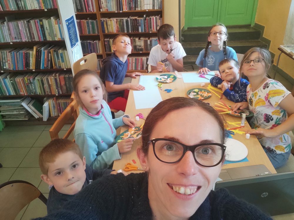Siedmioro dzieci oraz bibliotekarka pozują z uśmiechami do zdjęcia. Dzieci siedzą przy stole, na którym leżą kolorowe paski papieru i talerzyki papierowe.