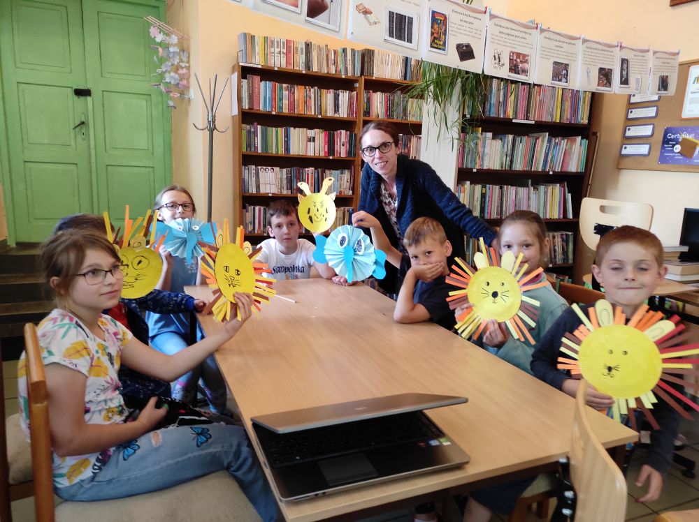 Siedmioro dzieci siedzi przy stole. Pięcioro z nich prezentuje wykonane własnoręcznie lwy z żółtych talerzyków papierowych i pomarańczowo-brązowych pasków papieru. Chłopiec i dziewczynka trzymają w rękach słonie z niebieskiego papieru.