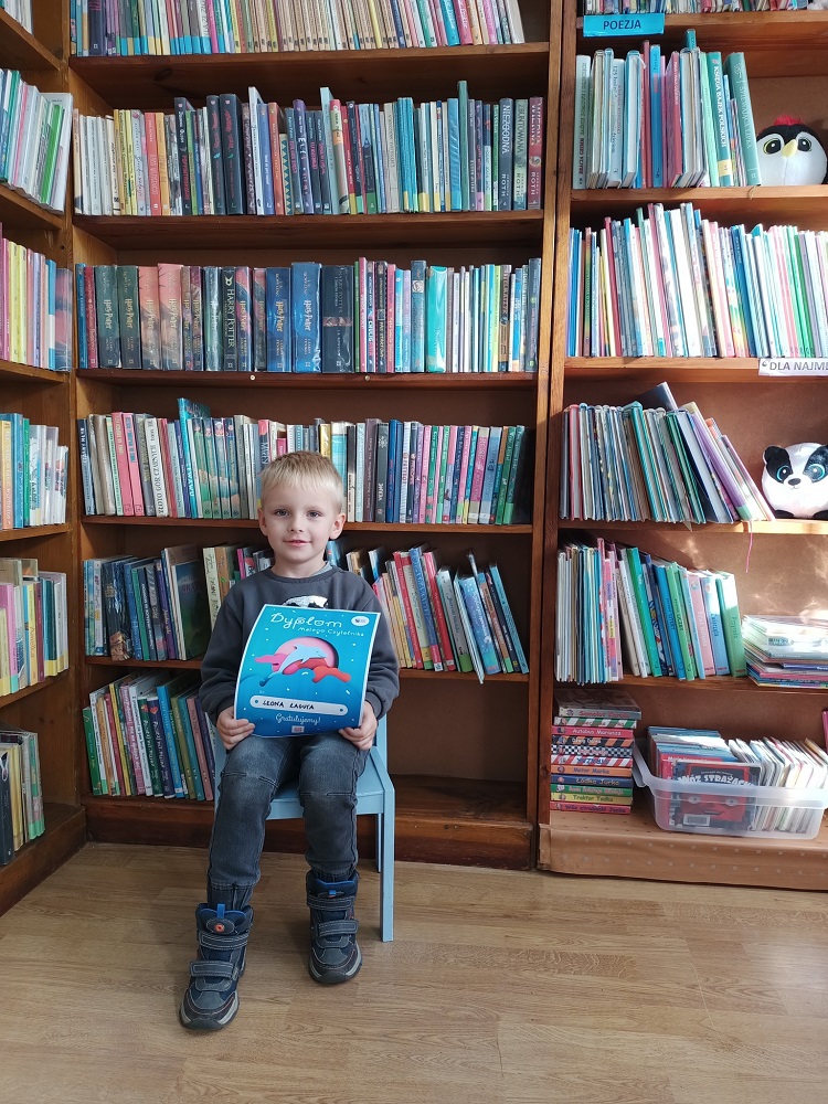 Siedzący mały chłopiec, trzymający dyplom. W tle regały z książkami.