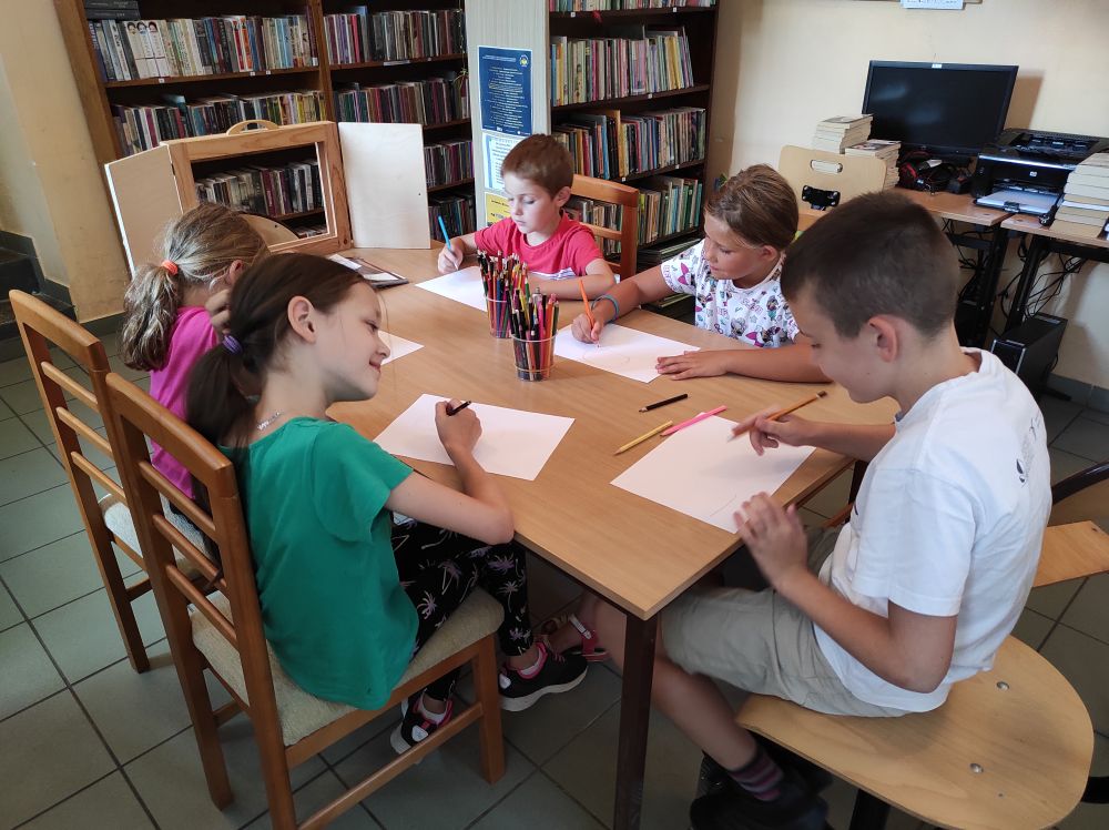 Pięcioro dzieci, trzy dziewczynki i dwóch chłopców, siedzi przy stole i malują kredkami.