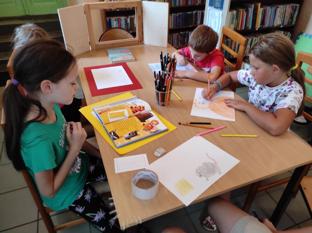 Pięcioro dzieci siedzi przy stole. Dwoje z nich maluje kredkami obrazki. Na stole stoją kubeczki z kolorowymi kredkami oraz leży otwarta książka.