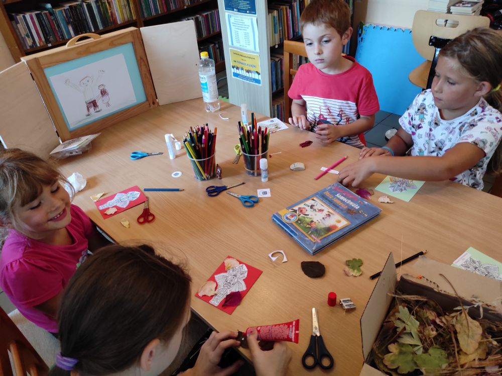 Czworo dzieci siedzi przy stole. Wykonują prace plastyczne. Na kolorowych kartkach naklejają suszone rośliny oraz wycięte postacie zwierząt.