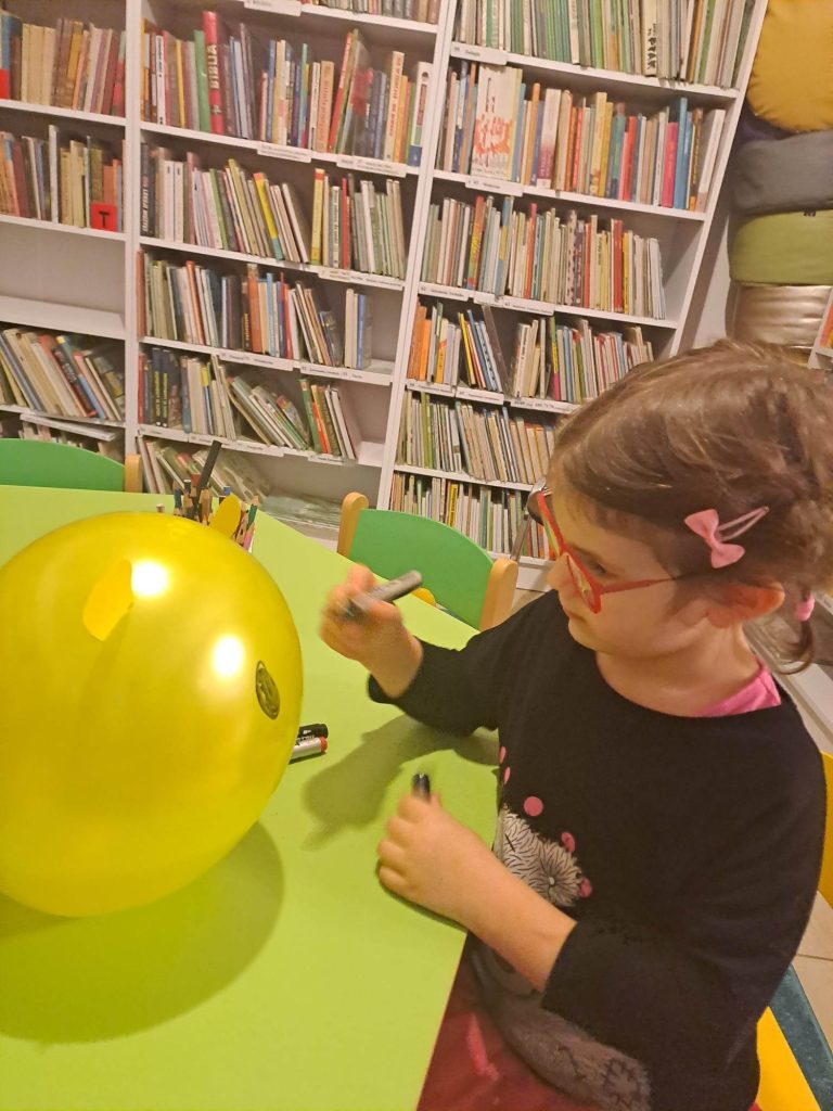 Siedząca przy stoliku dziewczynka mazakiem koloruje balona. W tle regały z książkami.