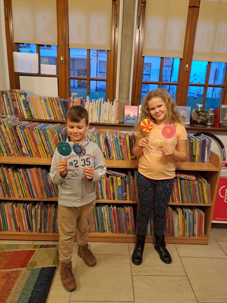 Na zdjęciu stoi dwoje dzieci. Trzymają w rękach własnoręcznie wykonane lizaki. W tle jest okno i regały z książkami.