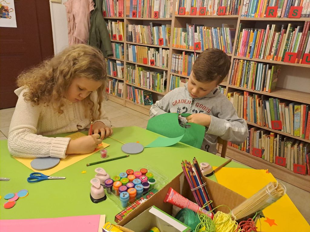 Dwoje dzieci siedzi przy zielonym stoliku na którym leżą materiały plastyczne: pieczątki, kleje, wycinanki, tasiemki. Za nimi znajdują się regały z dużą ilością książek.