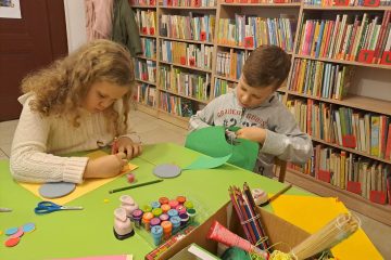 Dwoje dzieci siedzi przy zielonym stoliku na którym leżą materiały plastyczne: pieczątki, kleje, wycinanki, tasiemki. Za nimi znajdują się regały z dużą ilością książek.
