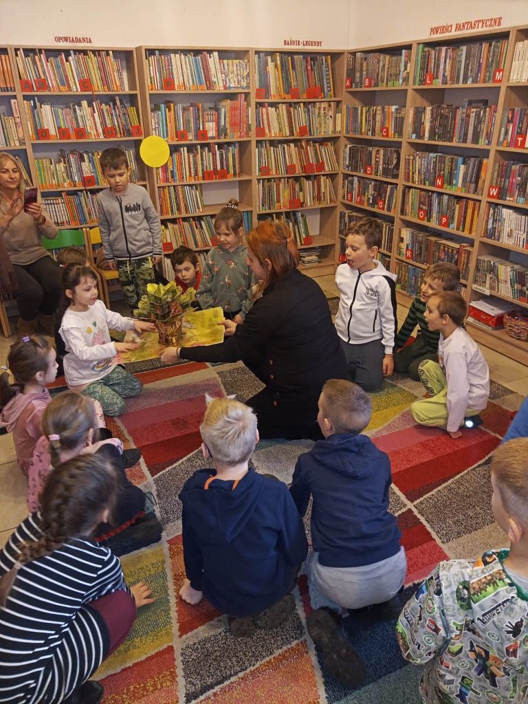 Na kolorowym dywanie siedzą w kółku dzieci. W środku pani bibliotekarka trzyma książkę. W tle regały z książkami.