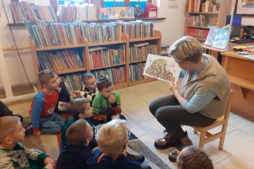 Po lewej stronie na kolorowym dywanie siedzą dzieci. Po prawej stronie na krzesełku siedzi pani bibliotekarka, która w rękach trzyma książkę. W tle regały z książkami, biurko, okna.