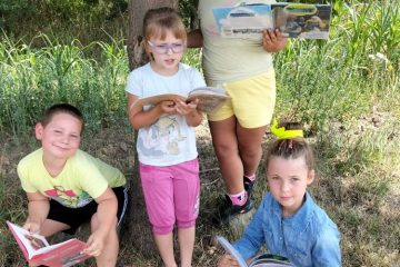 biblioteka w plenerze – dzieci czytają książki , chłopiec i dziewczyna siedzą na trawie a dwie dziewczynki stoją pod drzewem też z książkami
