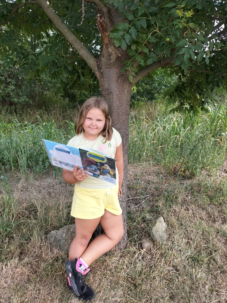 dziewczynka uśmiechnięta stoi pod drzewem i w prawej ręce trzyma otwartą książkę, w tle drzewa i trawa