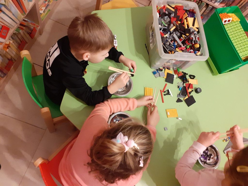 Na zdjęciu widać dzieci, które siedzą przy stole i pałeczkami przenoszą klocki lego do metalowych miseczek. W tle podłoga i półki z książkami.