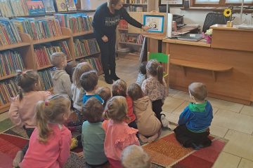 Na dywanie siedzi grupka dzieci, pani bibliotekarka pokazuje im teatrzyk. W tle biurko, okno, i półki z książkami.