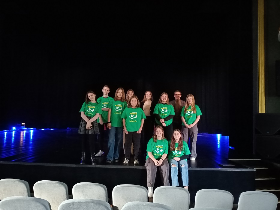11. Grupa osób w zielonych koszulkach stojąca na scenie. Przed nimi dwie osoby w zielonych koszulkach siedzące na scenie.