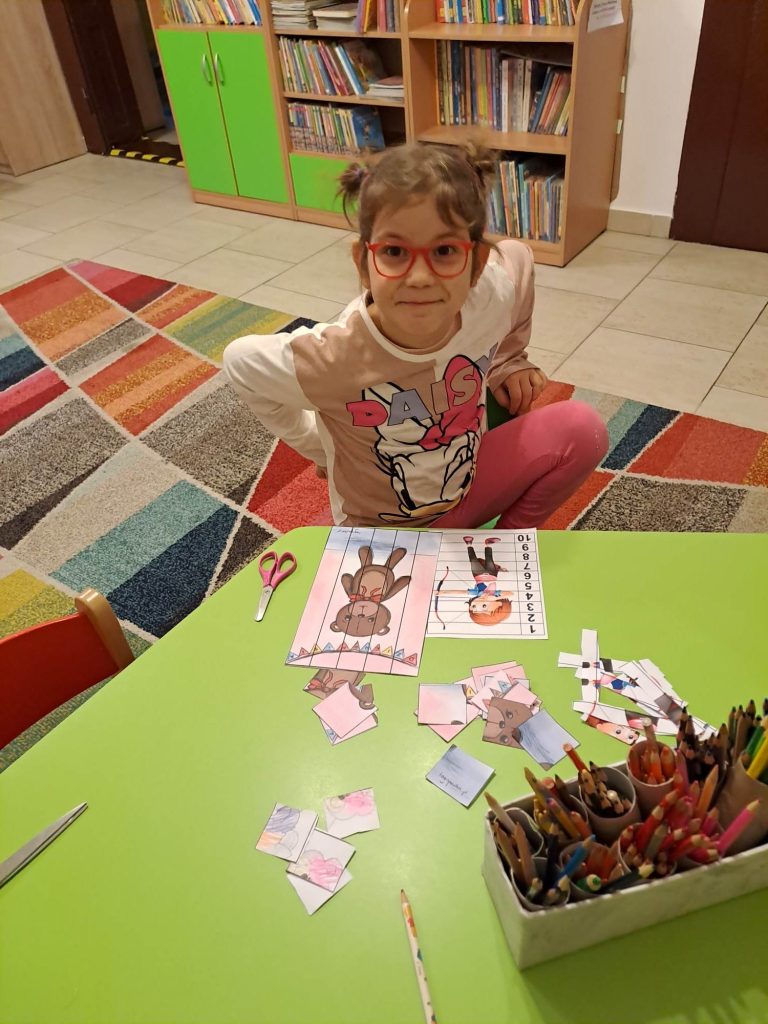 Przy stoliku siedzi dziewczynka i się uśmiecha, na stole znajdują się materiały plastyczne, w tle podłoga i regały z książkami.