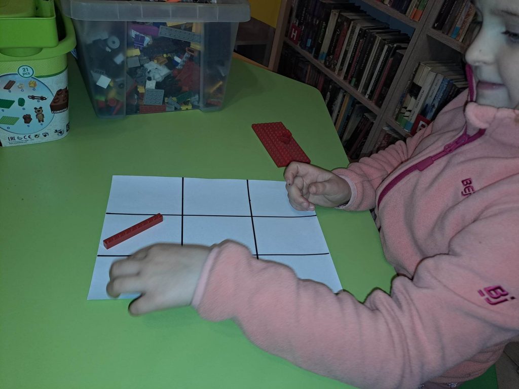 Przy stole siedzi dziewczynka, na planszy do gry w kółko-krzyżyk układa klocki lego, w tle regały z książkami.