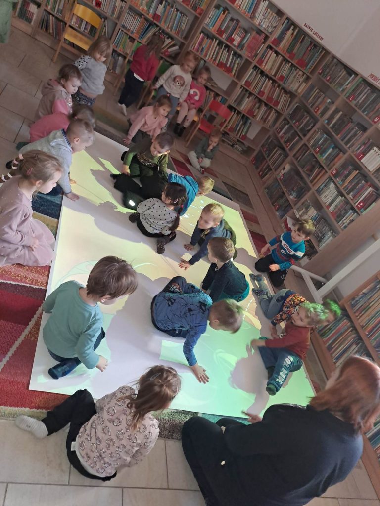 Dzieci bawią się na podłodze interaktywnej. W tle regały z książkami.