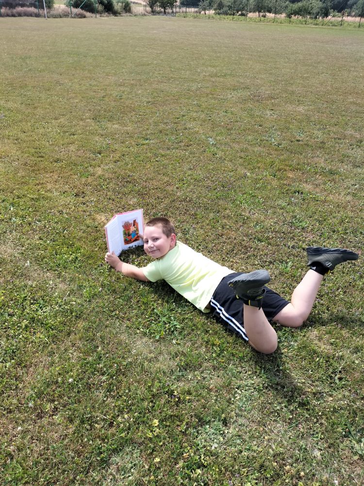 chłopiec leży na trawie z ugiętymi nogami do góry a w rękach trzyma opartą o trawę książkę