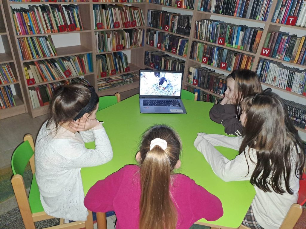 Dzieci siedzą przy stoliku i oglądają film na laptopie, który stoi na stole. W tle regały z książkami.