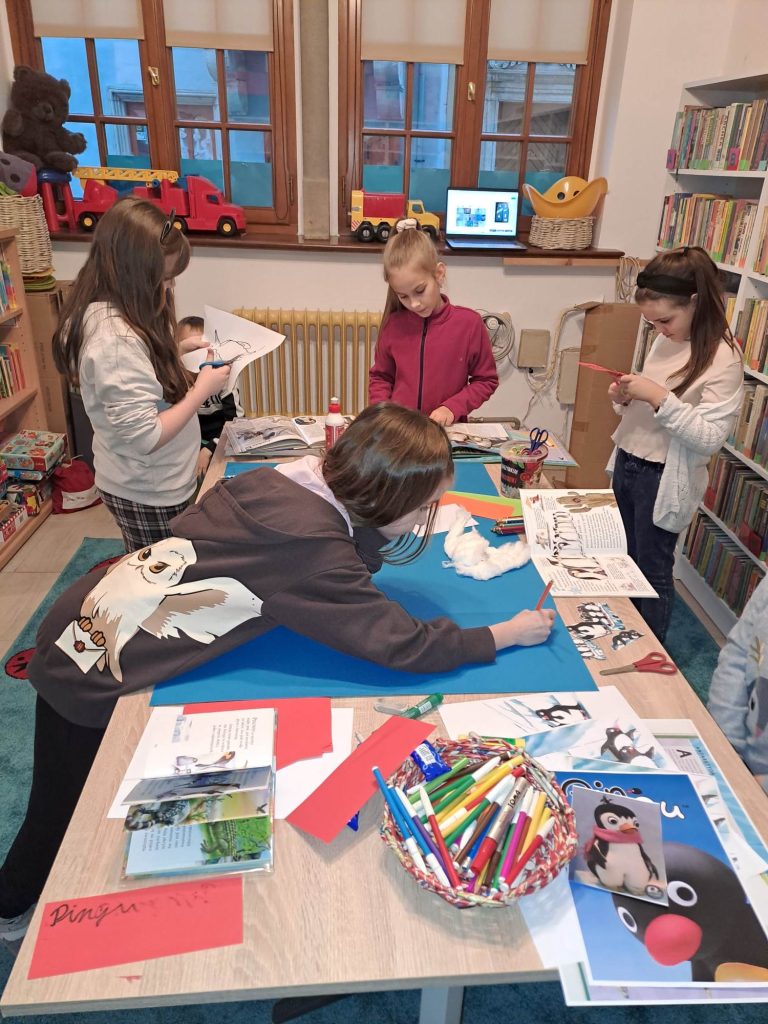Przy dużym stole dzieci robią lapbooka,na stole leżą materiały plastyczne, w tle okno ,zabawki i regały z książkami.