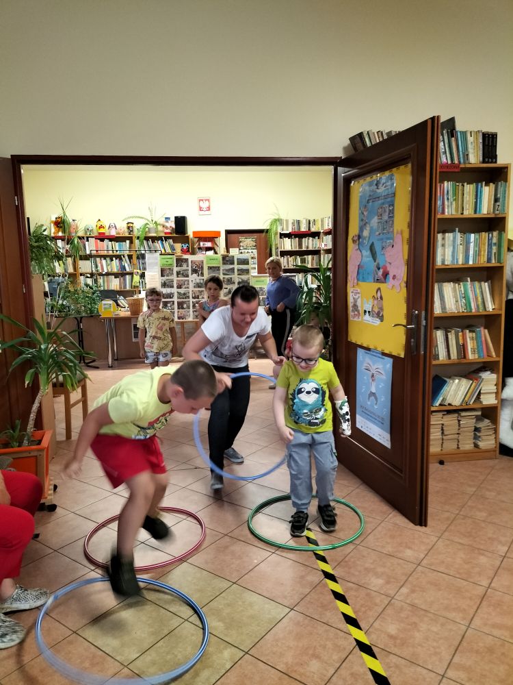 zabawa z hula-hop – dwóch chłopców i osoba dorosła przeskakują przez koła , w tle regały z książkami i troje dzieci kibicuje …