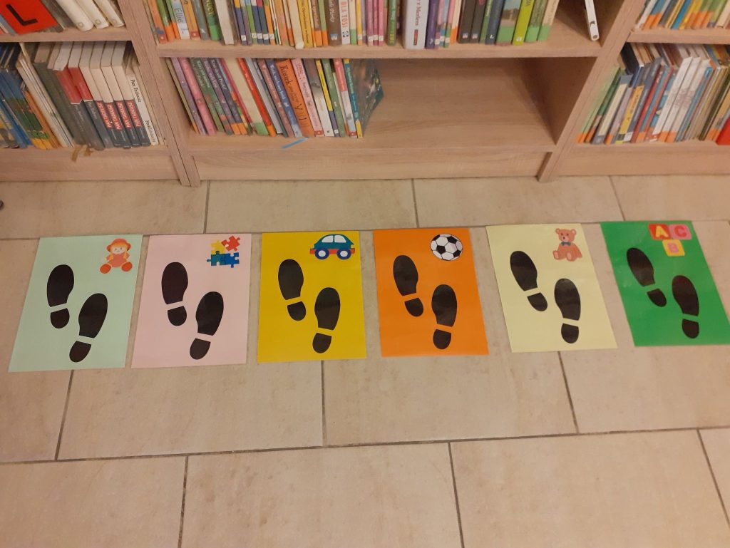 Na zdjęciu widać 6 kolorowych karteczek ze śladami obuwia i zabawkami, ułożonych na podłodze, w tle półki z książkami.