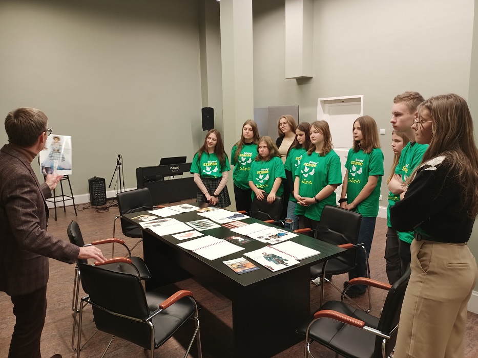 4. Od lewej stojący tyłem mężczyzna. Po środku stół z ilustracjami. Po prawej stojąca przodem grupa młodzieży w zielonych koszulkach z napisem „Równać szanse”.