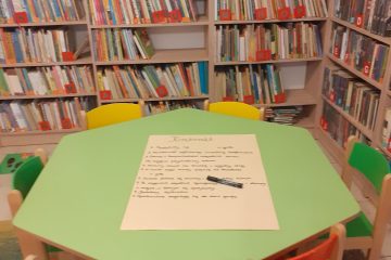 Na zielonym stole znajduje się kartka z zasadami Grupy Zabawowej, dookoła stolika stoją kolorowe krzesła, w tle regały z książkami i napis „Grupa Zabawowa”.