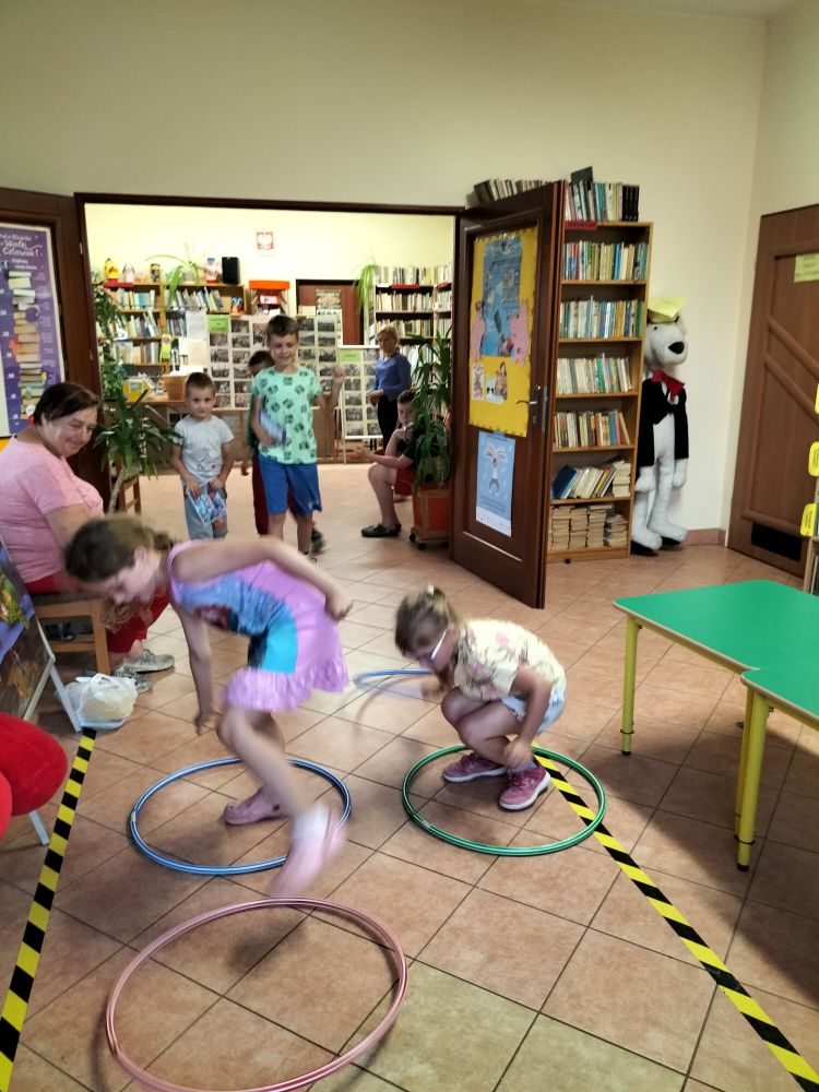 zabawa z hula-hop – dwie dziewczynki przeskakują przez koła, w tle regały z książkami i grupa dzieci kibicuje…