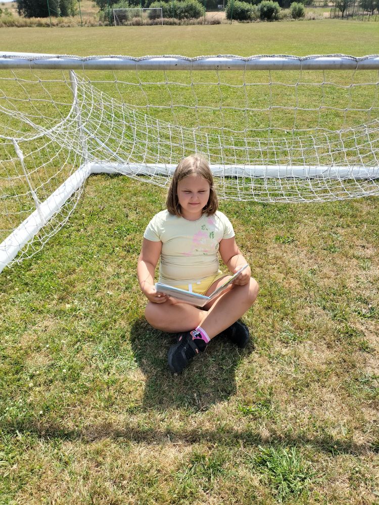 dziewczynka siedzi na trawie po turecku i czyta książkę w tle przewrócona bramka