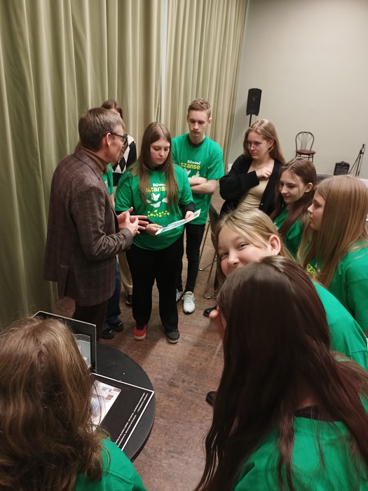 5. Grupa młodzieży w zielonych koszulkach słuchająca wykładu mężczyzny stojącego bokiem po lewej stronie.
