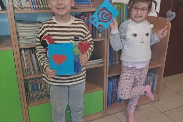 Na zdjęciu widać chłopca i dziewczynkę, którzy stoją i trzymają w rękach laurki walentynkowe, w tle półki z książkami.