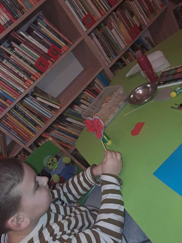 Przy zielonym stole siedzi chłopiec, w ręku trzyma serduszko na patyku. Na stole leżą materiały plastyczne, w tle regały z książkami.