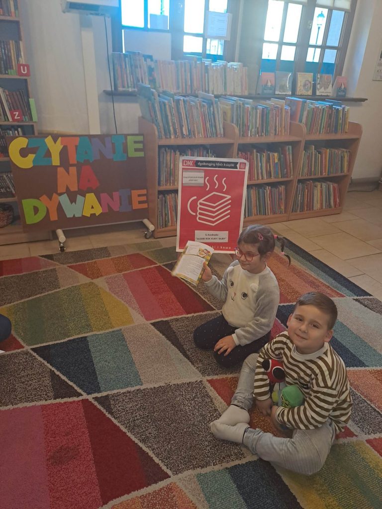 Na dywanie siedzą dzieci, w tle książki na półkach, plakat dkk i napis „Czytanie na dywanie”.