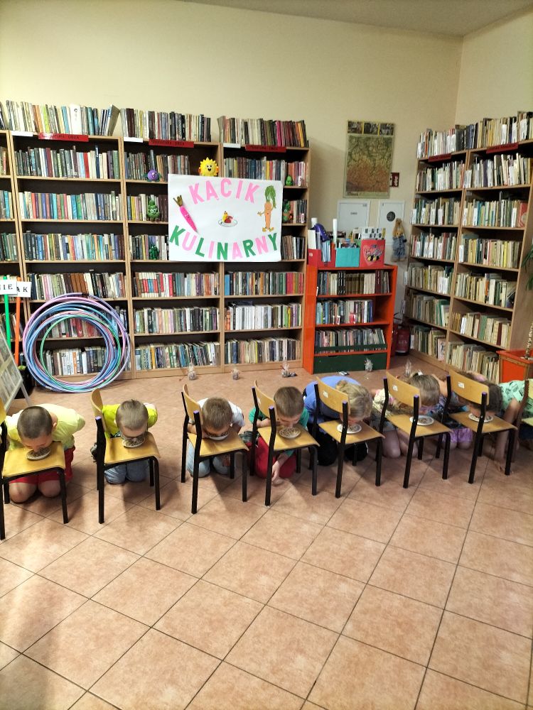 konkurs kto szybciej zje chrupki - ośmioro dzieci klęczy przy małych krzesełkach i zajada chrupki, w tle regały z książkami i plakat – kącik kulinarny …